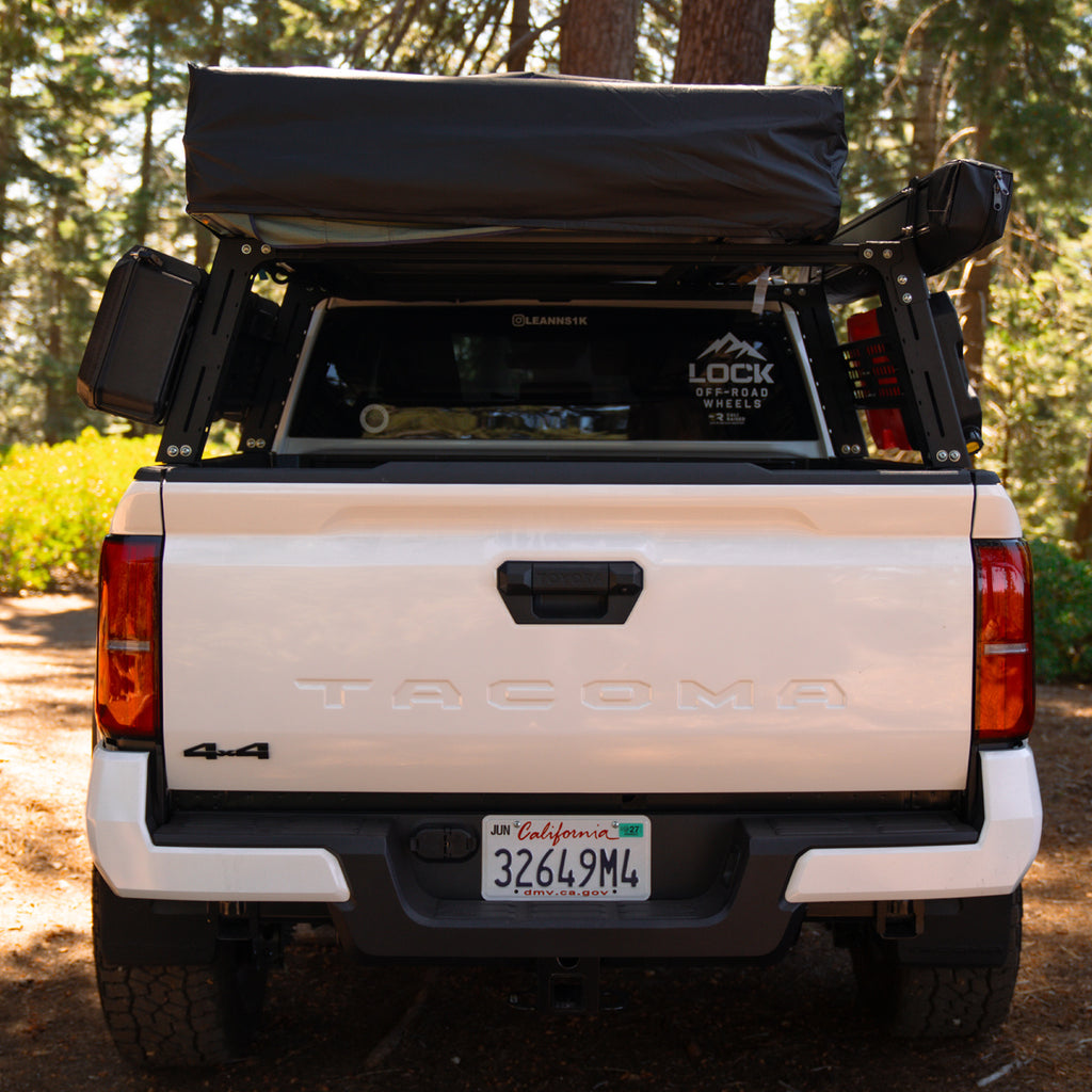 Rear view of a white Tacoma pickup truck in a forest setting, equipped with a black roof top tent.