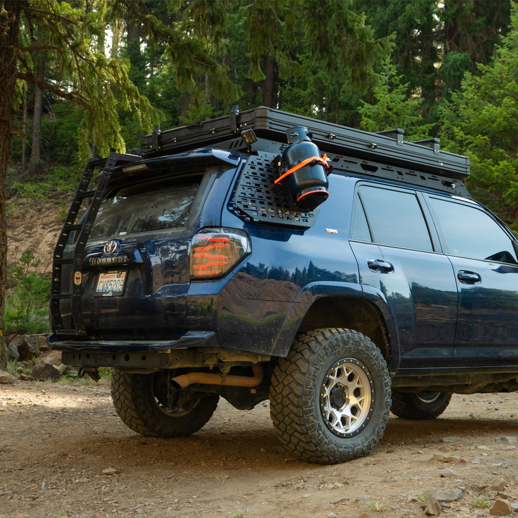 A dark blue off-road SUV parked on a dirt trail in a forest, showing a roof-mounted cargo box with a mounted cooler and a black rack system on the roof.