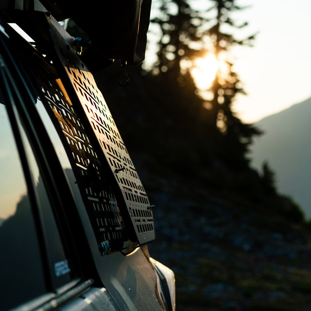 Close-up of a black metal roof rack with a slotted, vented design mounted on the side of a vehicle, set against a blurred mountain and pine forest background at sunset.