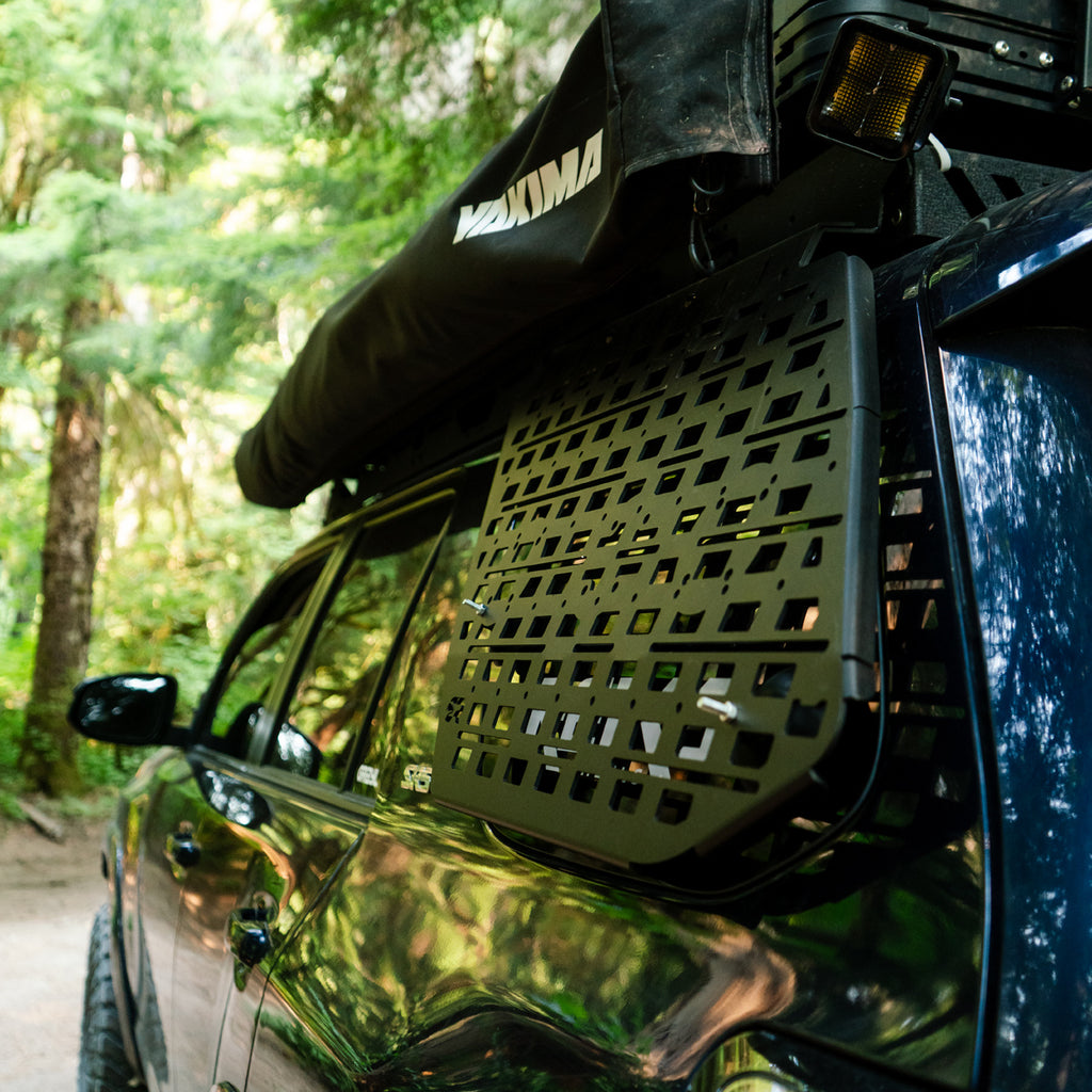 A black Maxxima roof tent and matching black storage rack mounted on the roof of a dark vehicle, photographed outdoors on a forest road.