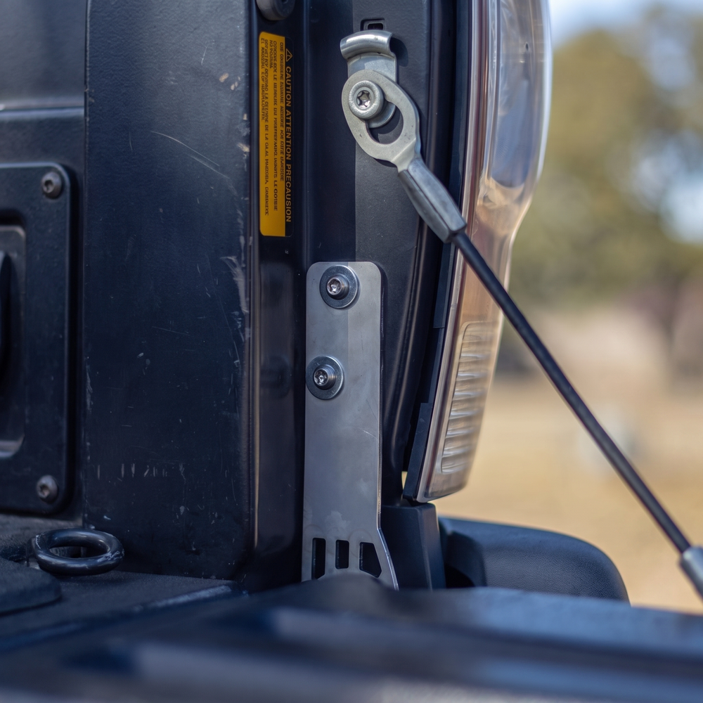 Cali Raised LED tailgate theft protector installed at the passenger-side tailgate hinge of a 2005-2015 Toyota Tacoma, secured with two supplied security bolts. Current protector shown.