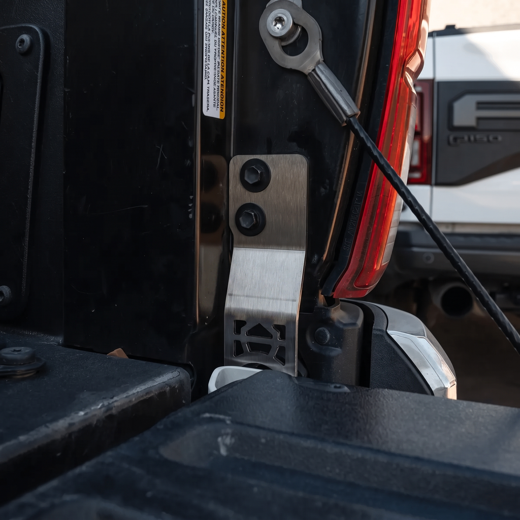 Close-up of a brushed metal tailgate latch bracket installed on the rear hatch of a black vehicle, with part of a red taillight and a white vehicle visible in the background.