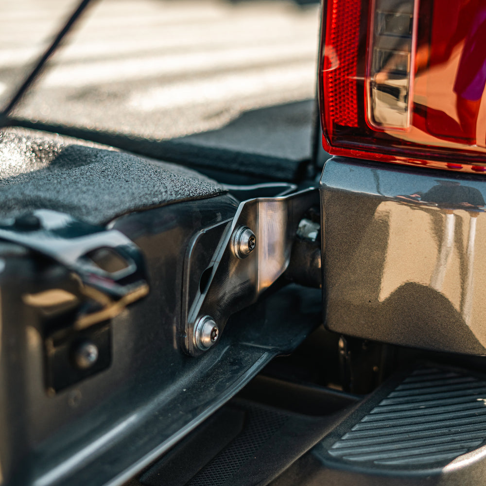 Cali Raised LED tailgate theft protector installed at the passenger-side tailgate hinge of a 2015-2026 Ford F-150, secured with two supplied security bolts. Current protector shown.