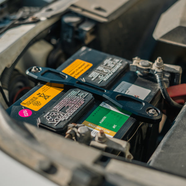A close-up of a car battery inside an open engine bay, with a battery clamp hold down resting across the battery and warning labels visible.