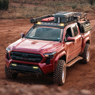 2024 Tacoma Roof Rack, Red 2024 Toyota Tacoma overlanding on a dirt trail, equipped with all-terrain tires, a front bumper light bar, and a fully loaded roof rack and bed rack