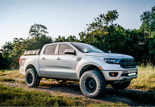 A lifted silver 2019-2024 Ford Ranger FX4 off-road pickup equipped with a Cali Raised LED Overland Bed Rack, parked on a dirt trail surrounded by trees at golden hour