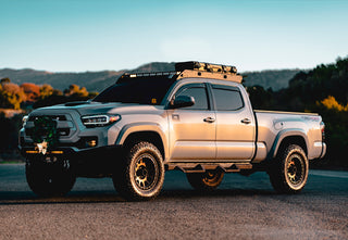 A silver Toyota Tacoma TRD Off-Road with a roof rack, mounted gear, and a full lighting setup, photographed during golden hour.