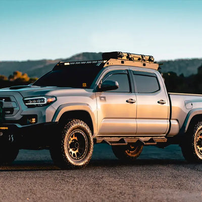 A silver Toyota Tacoma TRD Off-Road with a roof rack, mounted gear, and a full lighting setup, photographed during golden hour.